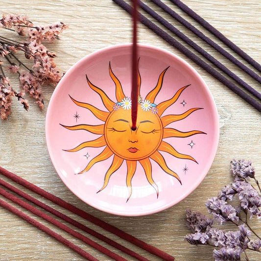 Pink plate with sun design, incense sticks, and flowers on a wooden surface